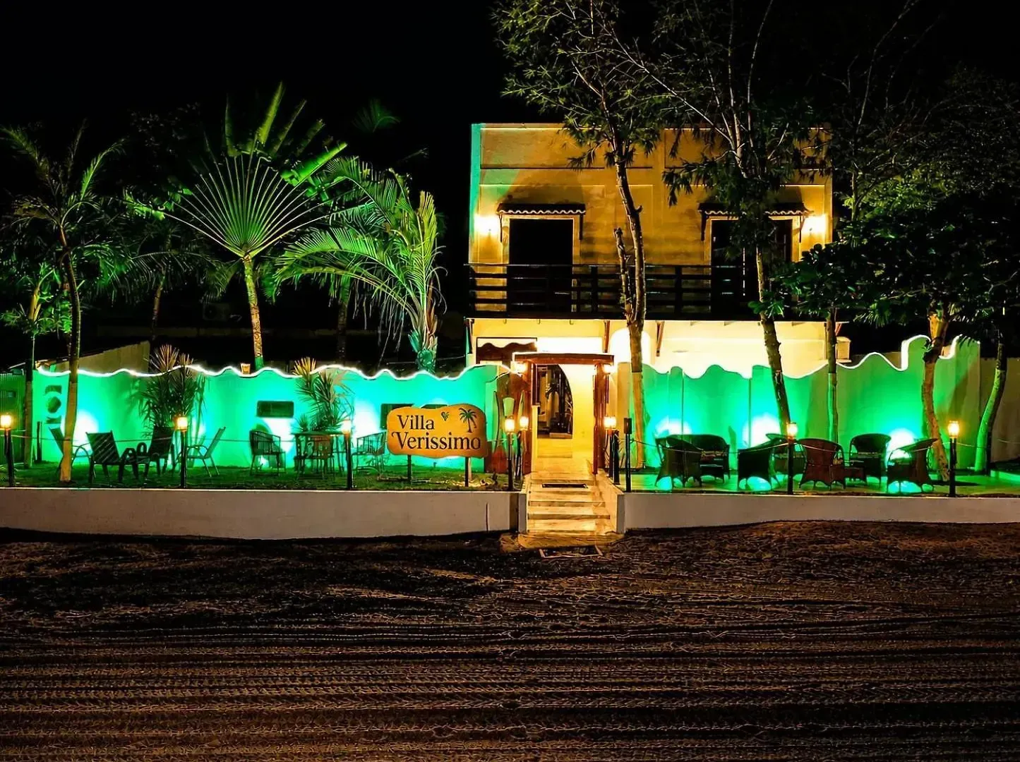 Vista noturna de um edifício bem iluminado com luzes verdes, palmeiras e uma entrada central com escadaria.