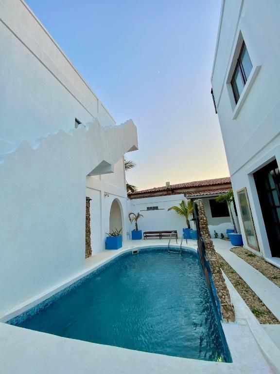 Piscina estreita e azul em um pátio de paredes brancas com espreguiçadeira e pérgola.