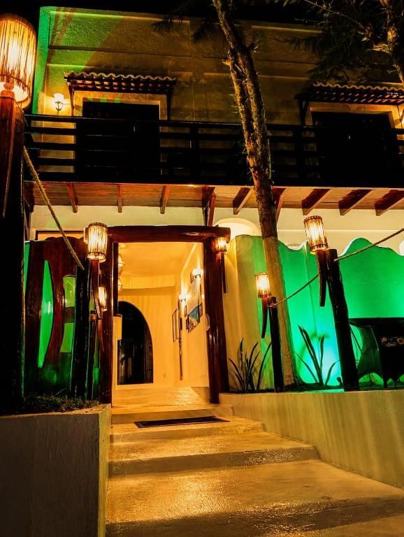 Entrada de um prédio de dois andares com iluminação noturna, luzes verdes ascendentes e uma porta com brilho aconchegante.