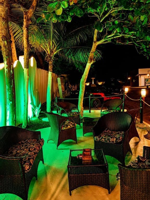Área de estar externa com cadeiras e mesas de vime, iluminada por luzes verdes ao lado de palmeiras à noite.