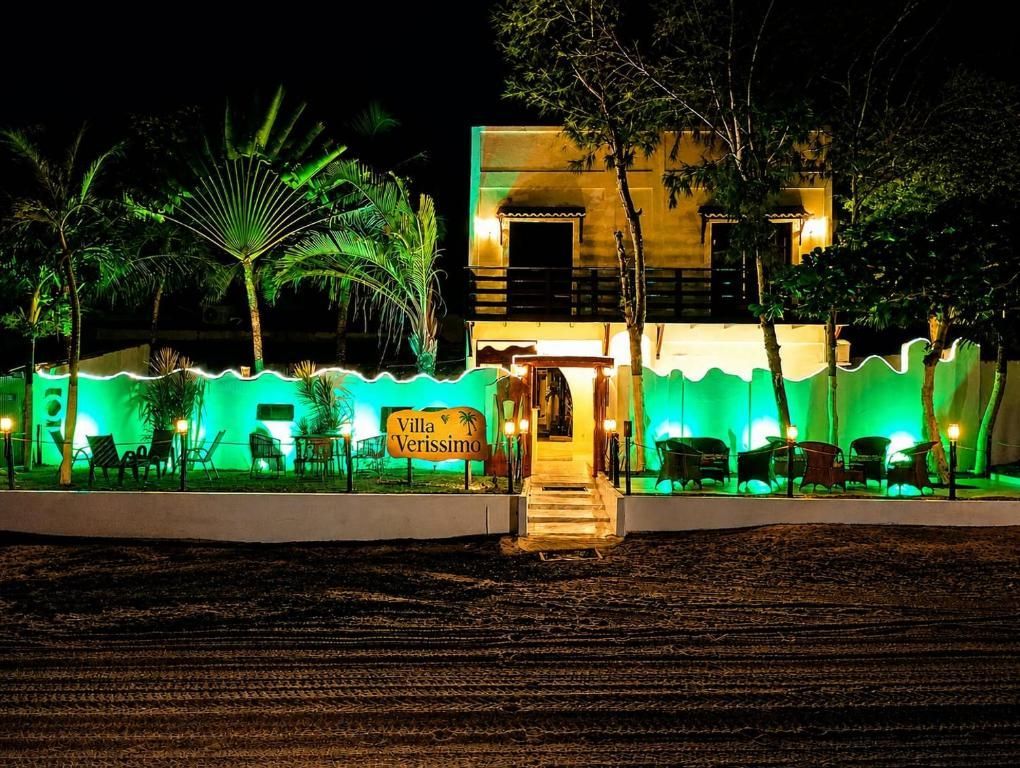 Vista noturna de um pequeno edifício iluminado com luzes verdes, palmeiras e uma escadaria central.