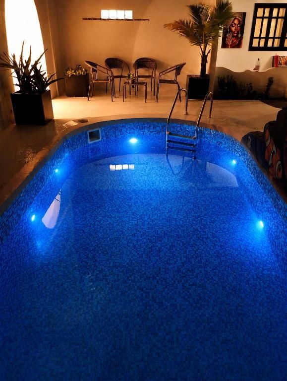 Piscina iluminada de azul à noite, ao lado de um pátio com cadeiras e vasos de plantas.