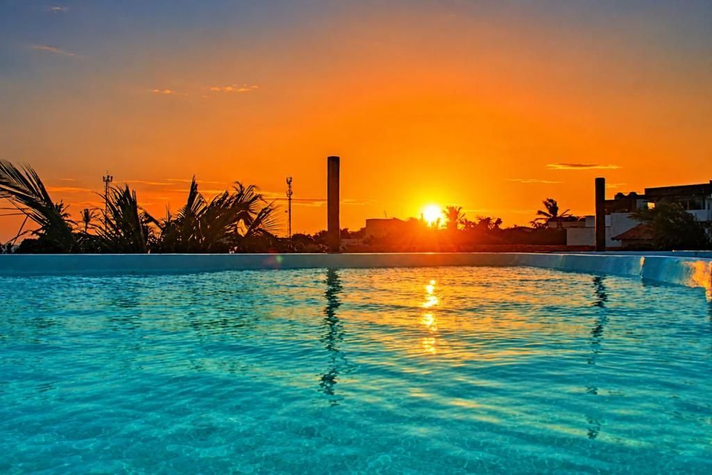 Pôr do sol sobre uma piscina turquesa com céu alaranjado e silhuetas de palmeiras.