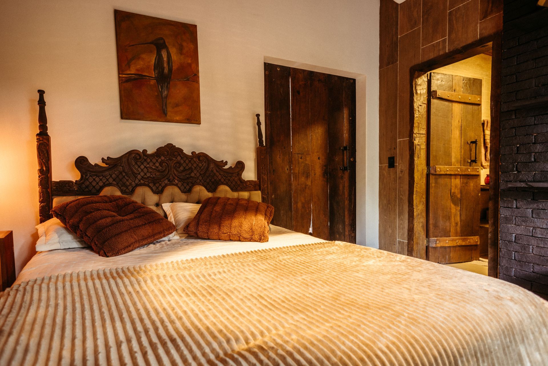 Quarto aconchegante com cama de madeira entalhada, colcha listrada, iluminação quente e porta aberta para um banheiro revestido com azulejos.