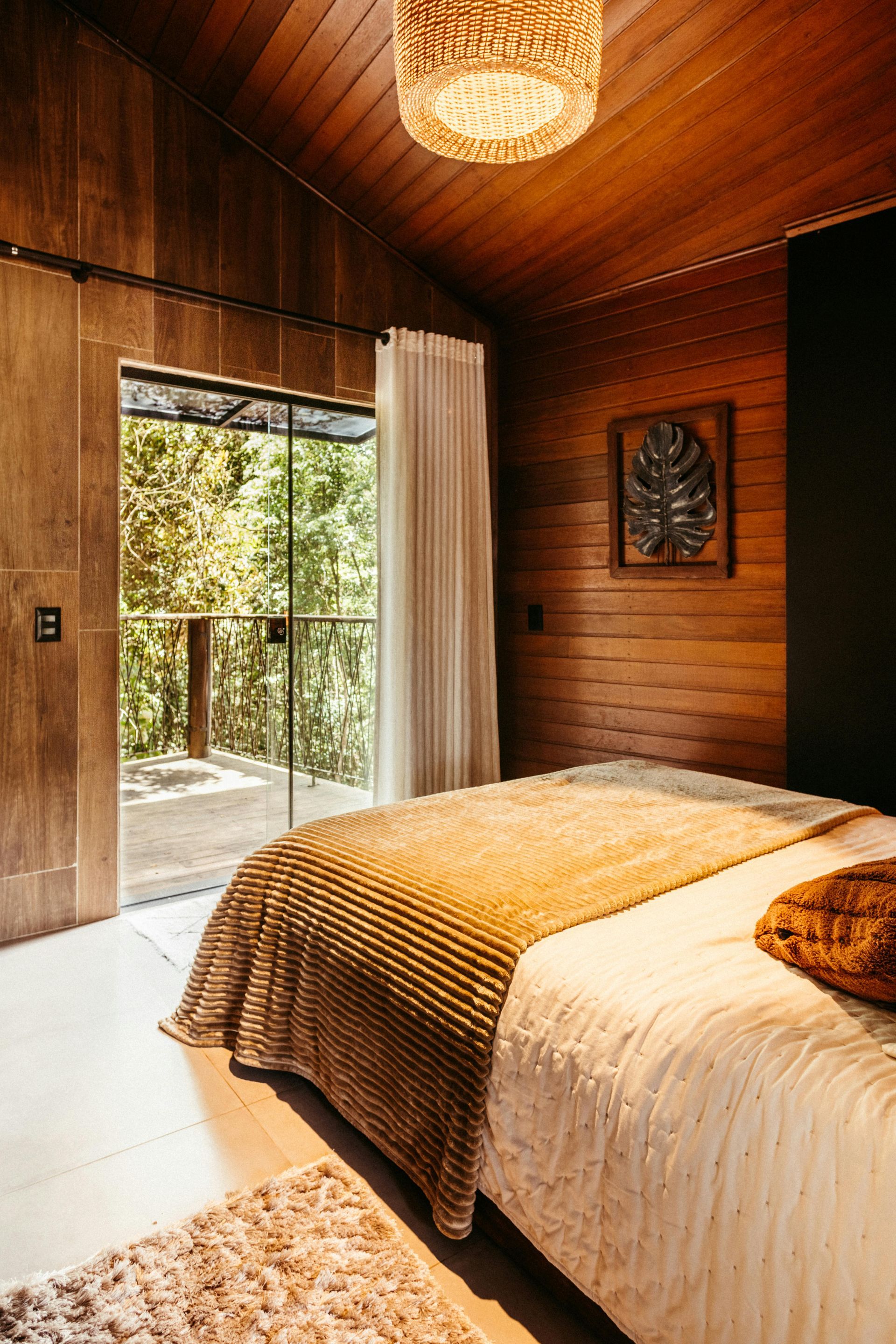 Quarto aconchegante de madeira com cama, luminária de vime e porta aberta para uma varanda arborizada com vista para a vegetação.