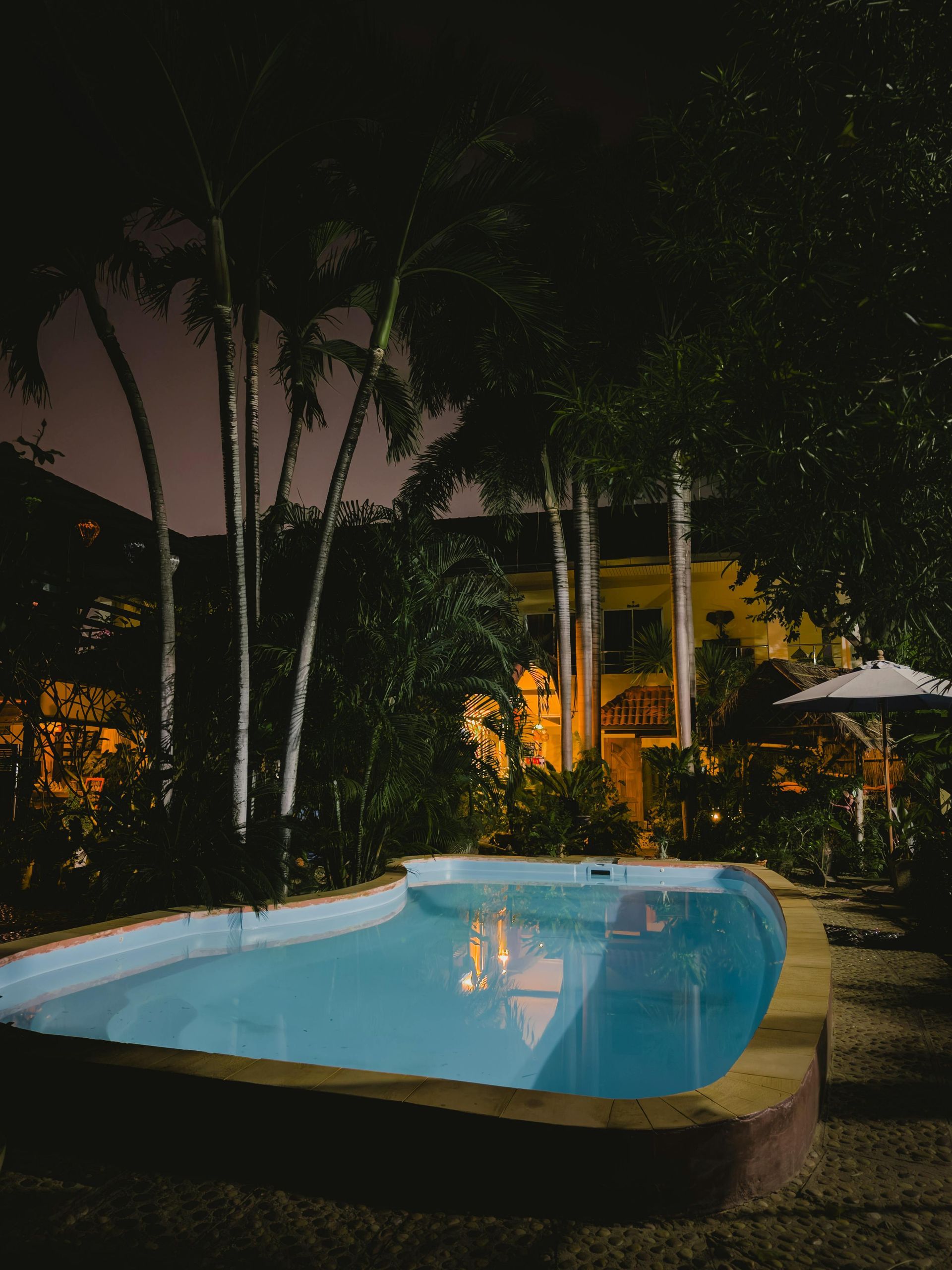 Piscina noturna com água azul, palmeiras e luzes aconchegantes em um pátio tropical.
