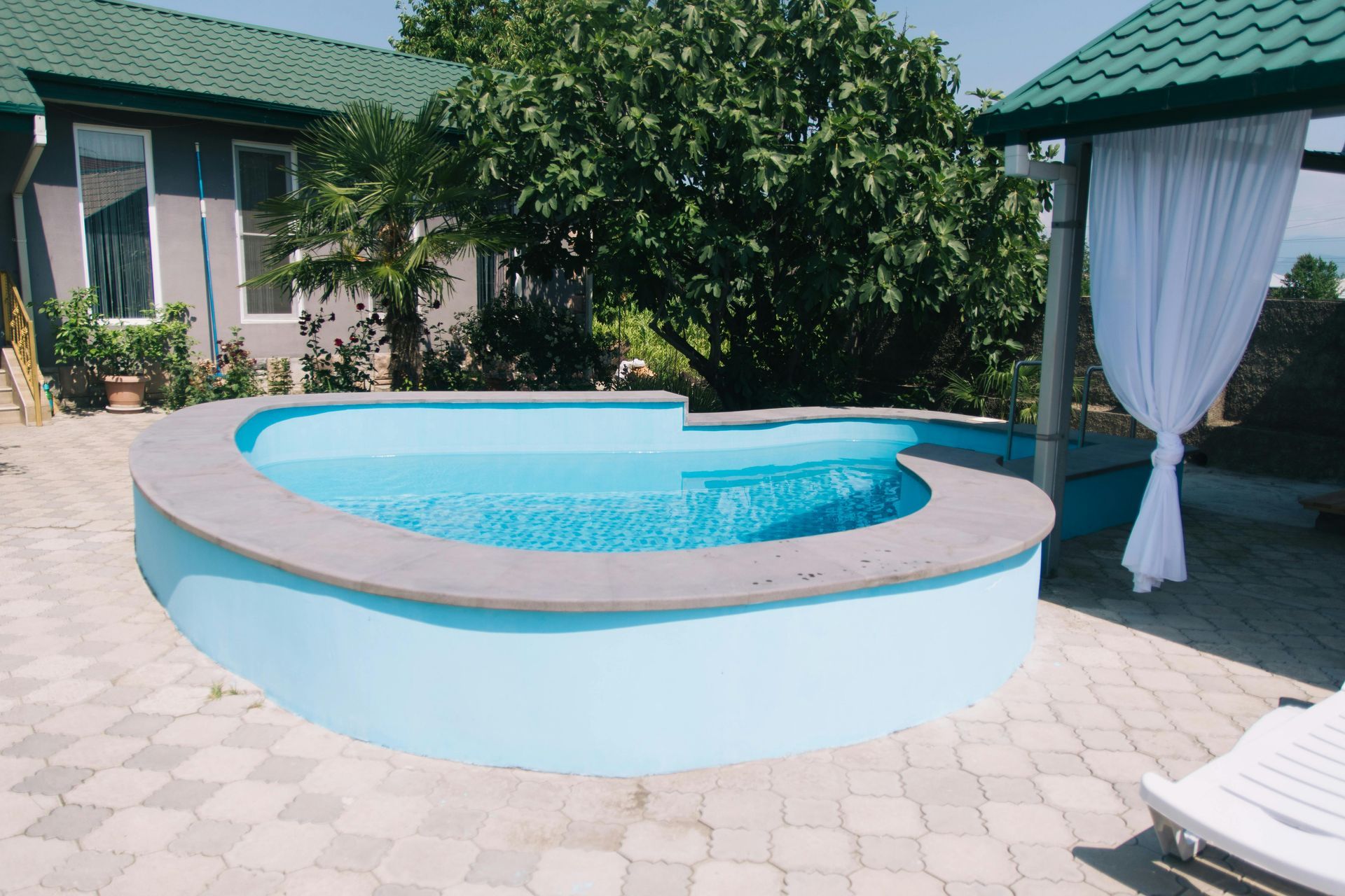 Piscina no quintal com água azul, pátio revestido com azulejos, cortinas de proteção e espreguiçadeira ao sol.