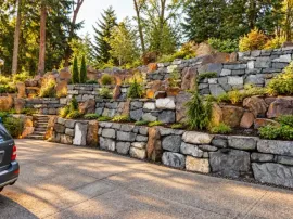 Construction worker in hard hat and safety vest building a gray block retaining wall beside a gravel road.