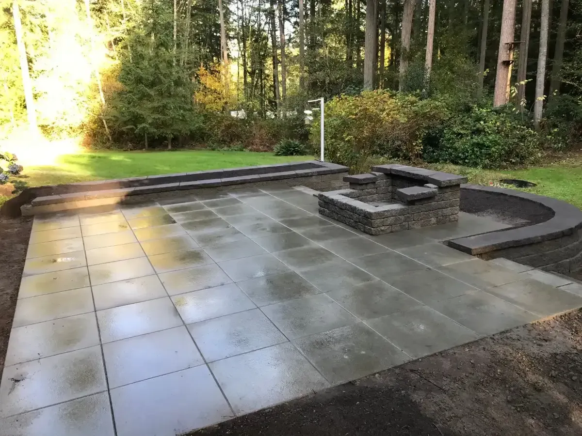 Sunny courtyard patio with tan stone tiles, lush greenery, and a stone fountain beside a wall