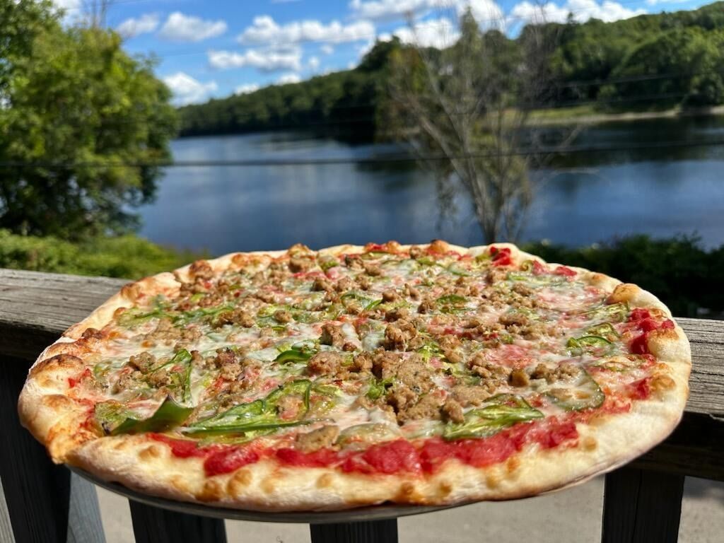 Pizza on a metal peel, in front of a wood-fired oven with flames.