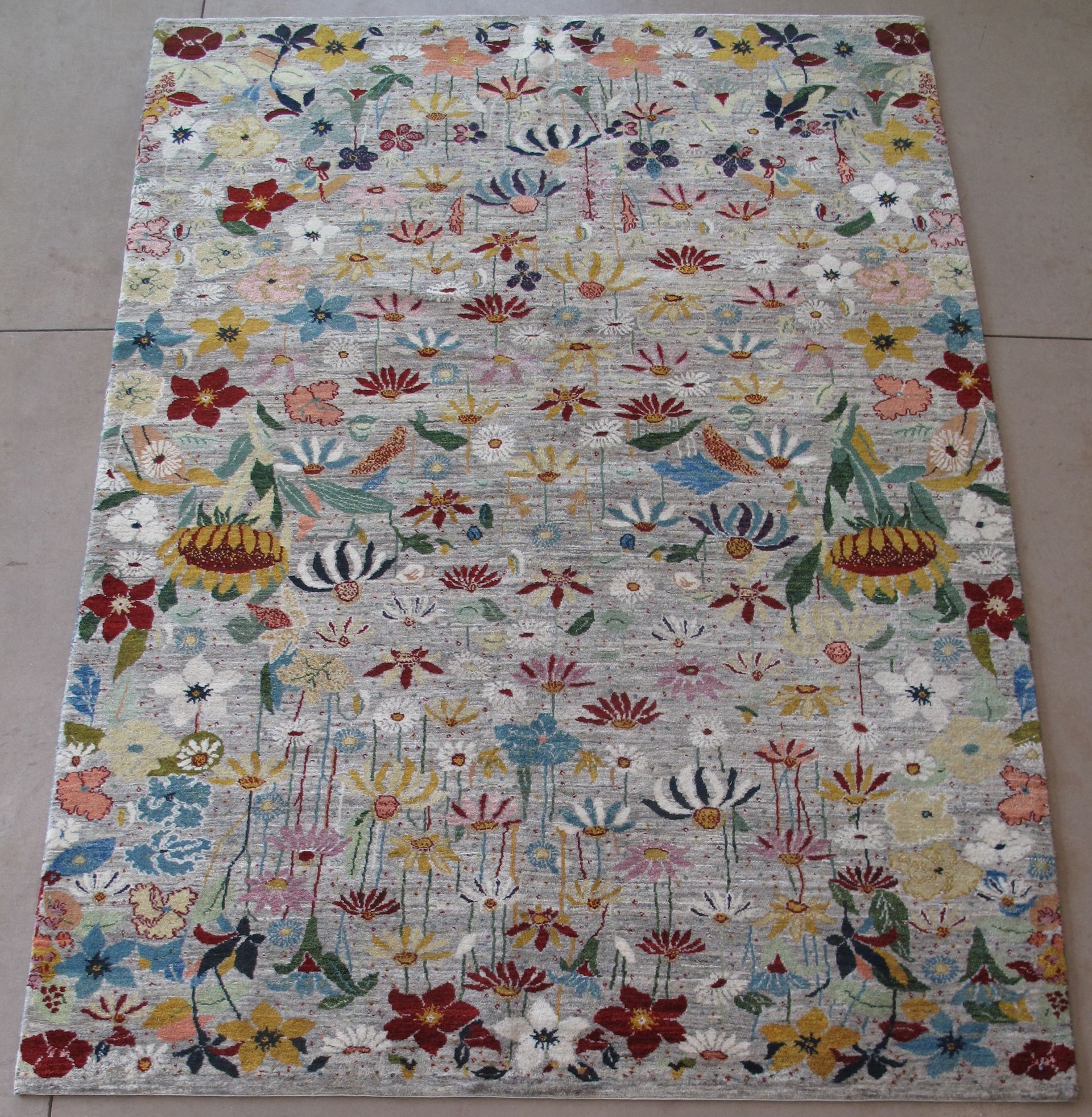 A rectangular area rug with a light gray background, densely patterned with a colorful variety of flowers and foliage.