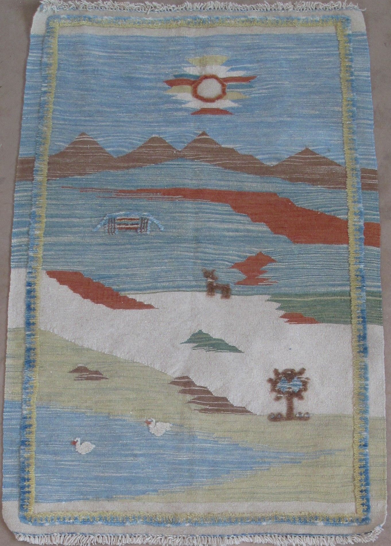 A rectangular rug featuring a landscape with a blue sky, mountains, a reddish landscape feature, a tree, and a pond.