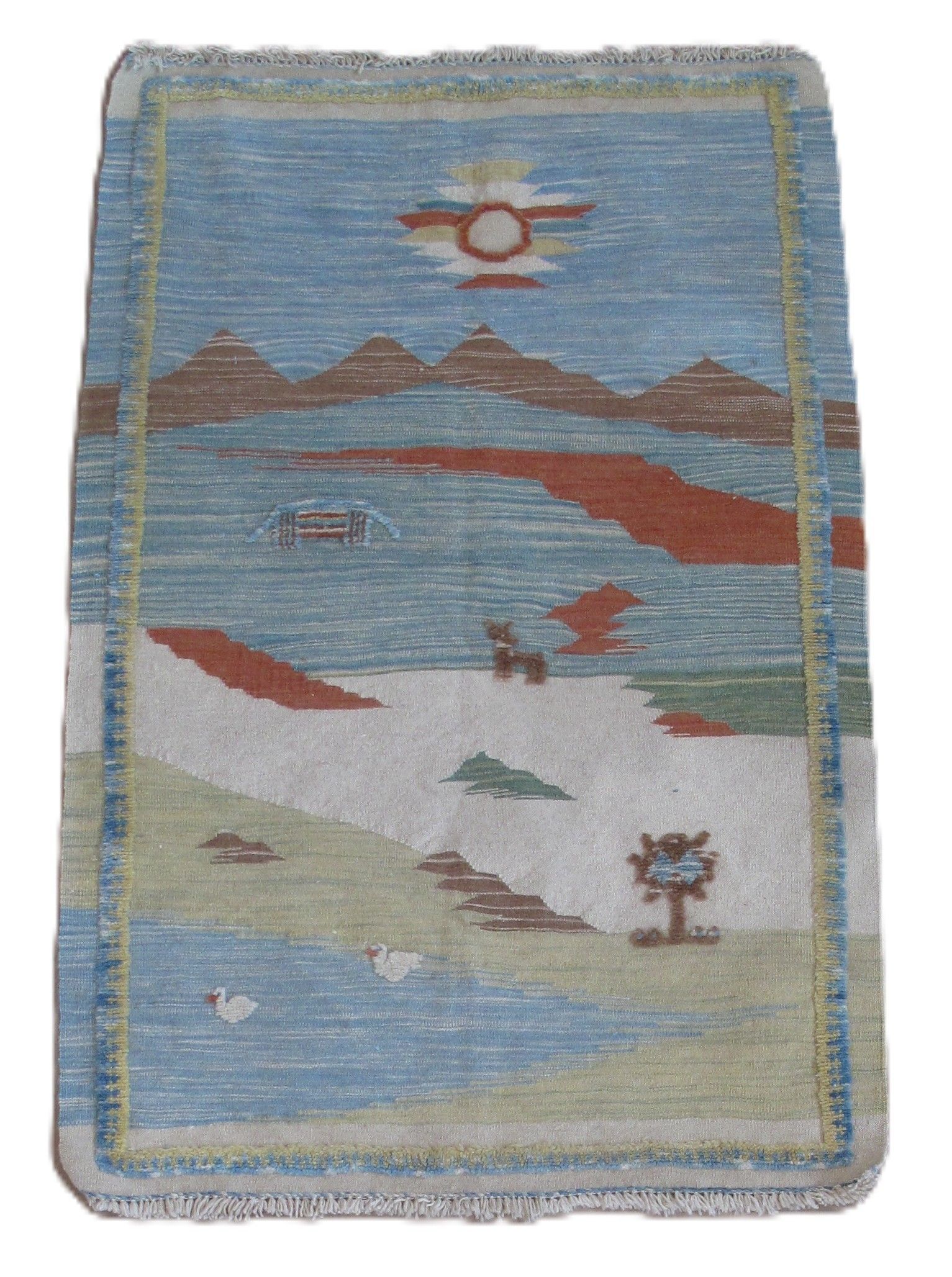 A rectangular rug featuring a landscape with a blue sky, mountains, a reddish landscape feature, a tree, and a pond.