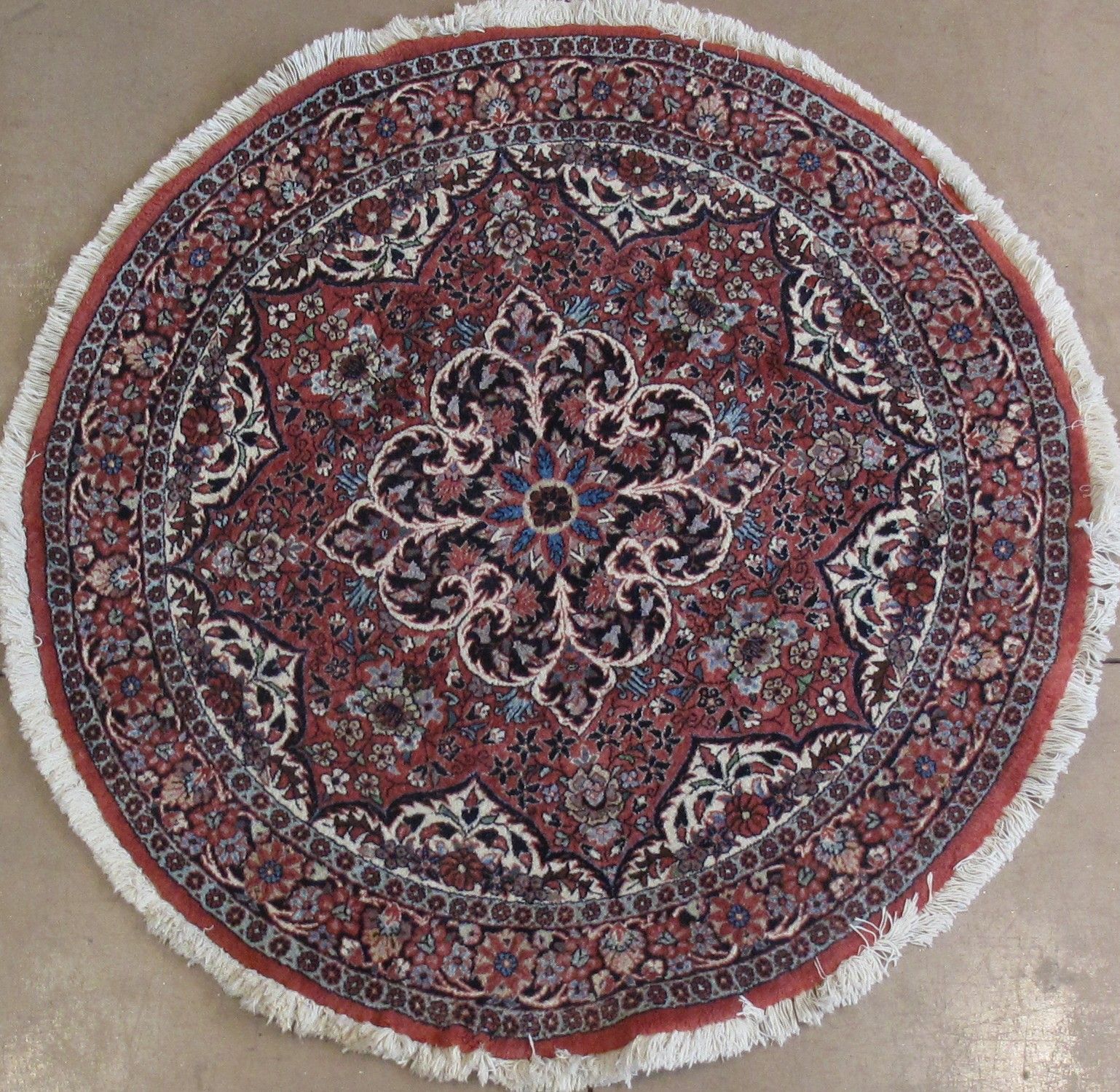 A round Persian-style rug with an intricate floral medallion pattern in red, blue, cream, and black, featuring white fringe.