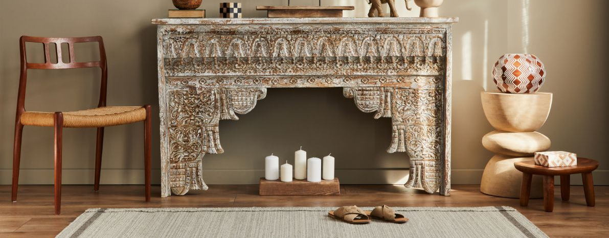 An ornate, carved wood fireplace mantel centerpiece sits against a wall, flanked by a wooden chair, rug, and stools.