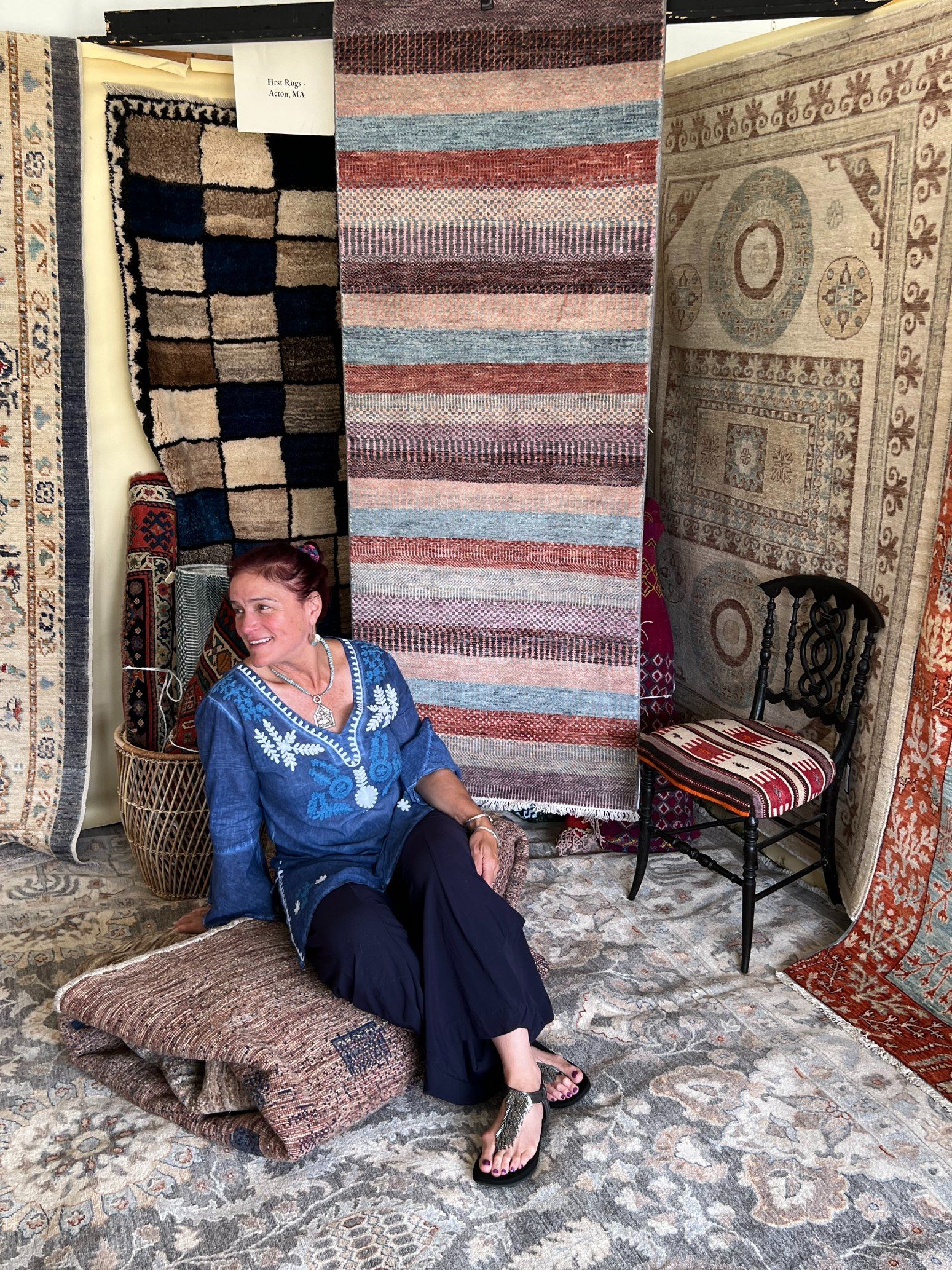 Cynthia First sitting in a rug shop surrounded by hanging patterned carpets and textiles