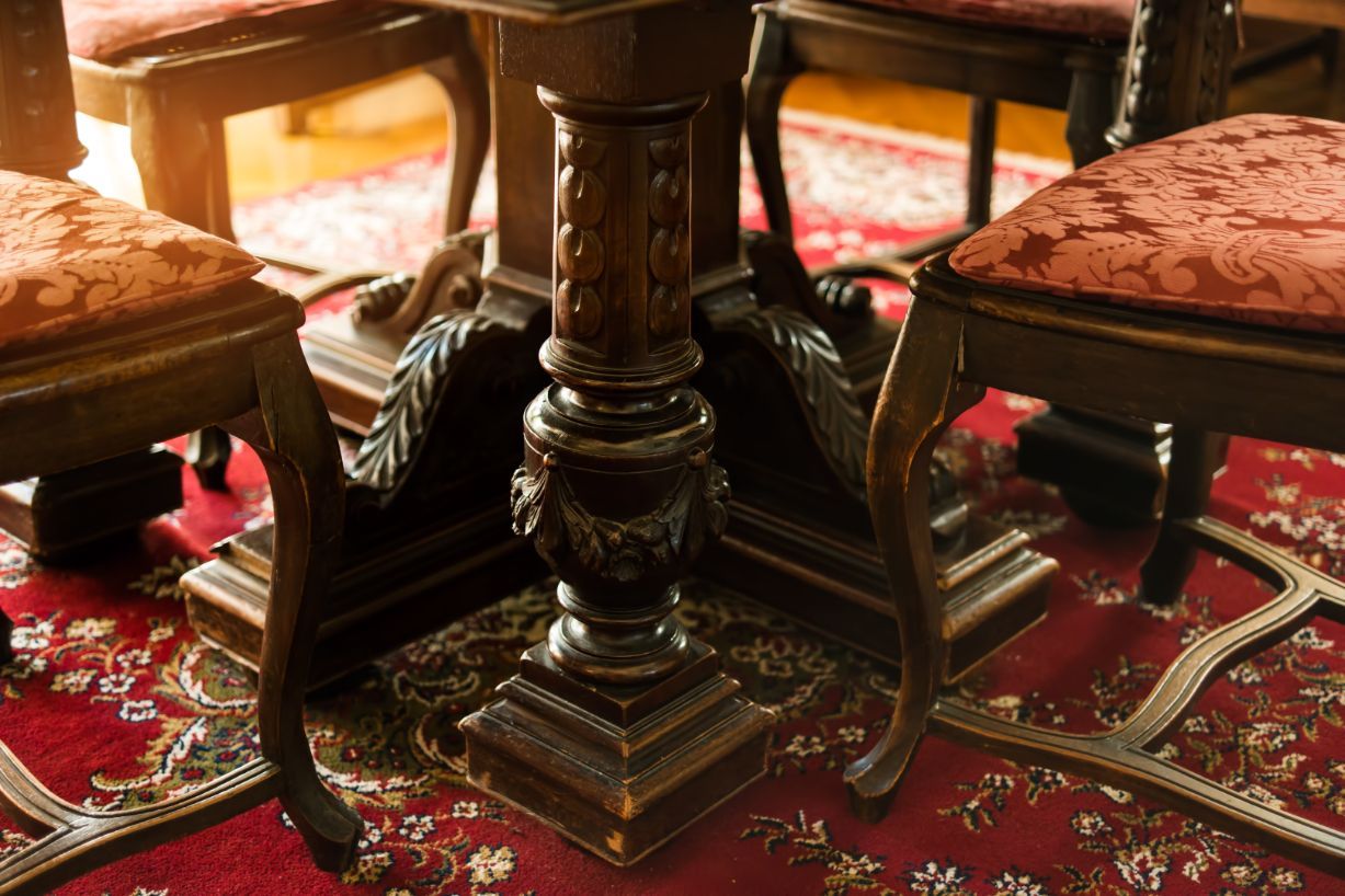 Close-up of a dark wooden dining table base with ornately carved details, surrounded by chairs and patterned red antique rug.