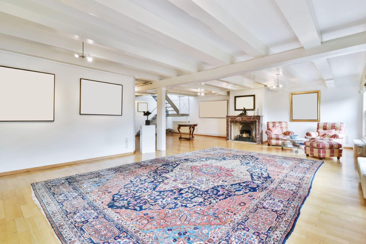 Spacious room with a large patterned rug, white walls, and wood beams.  Empty frames and a fireplace are also visible.