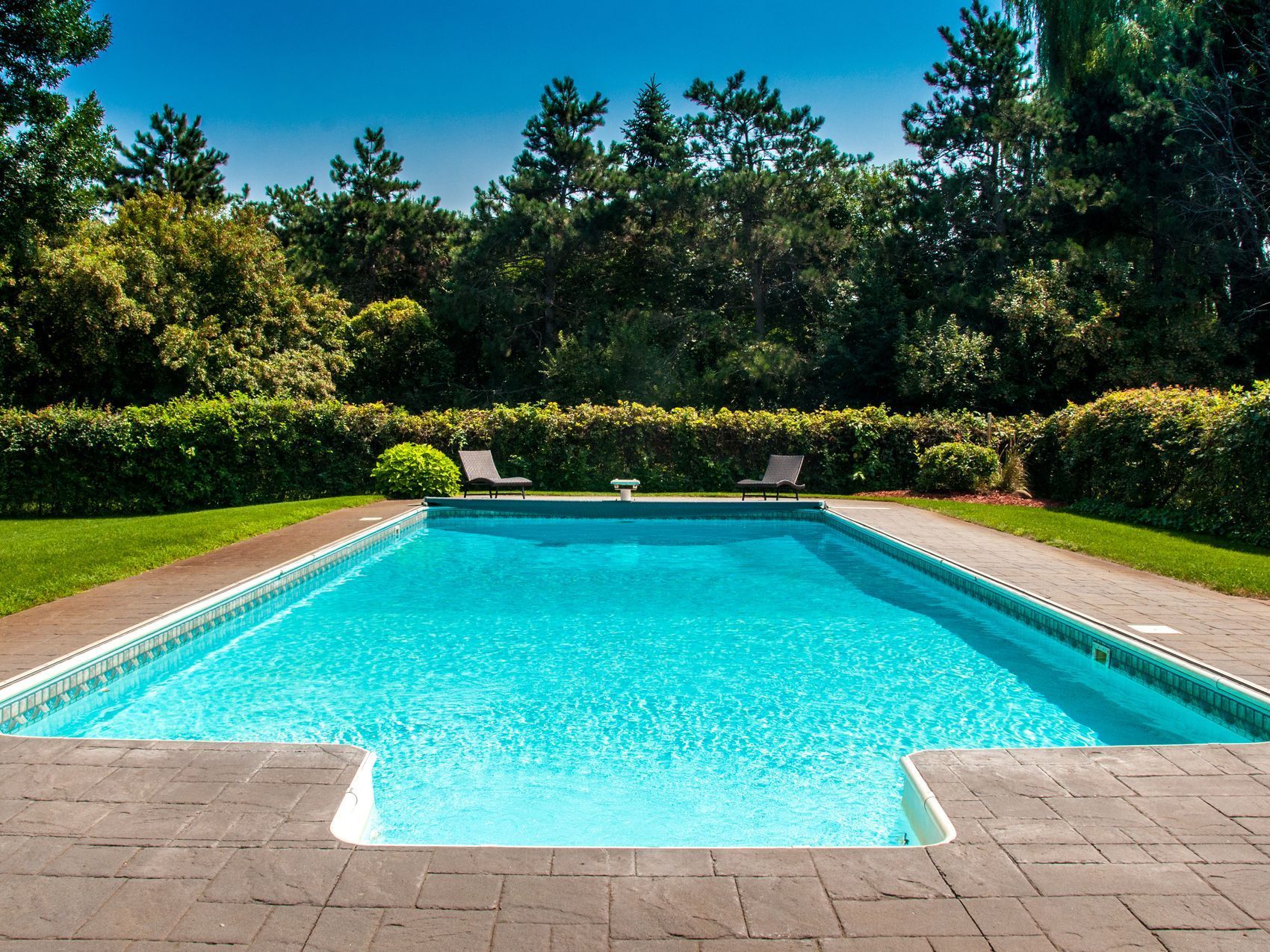 A clear blue swimming pool with brick surround; green lawn, and trees under a blue sky.