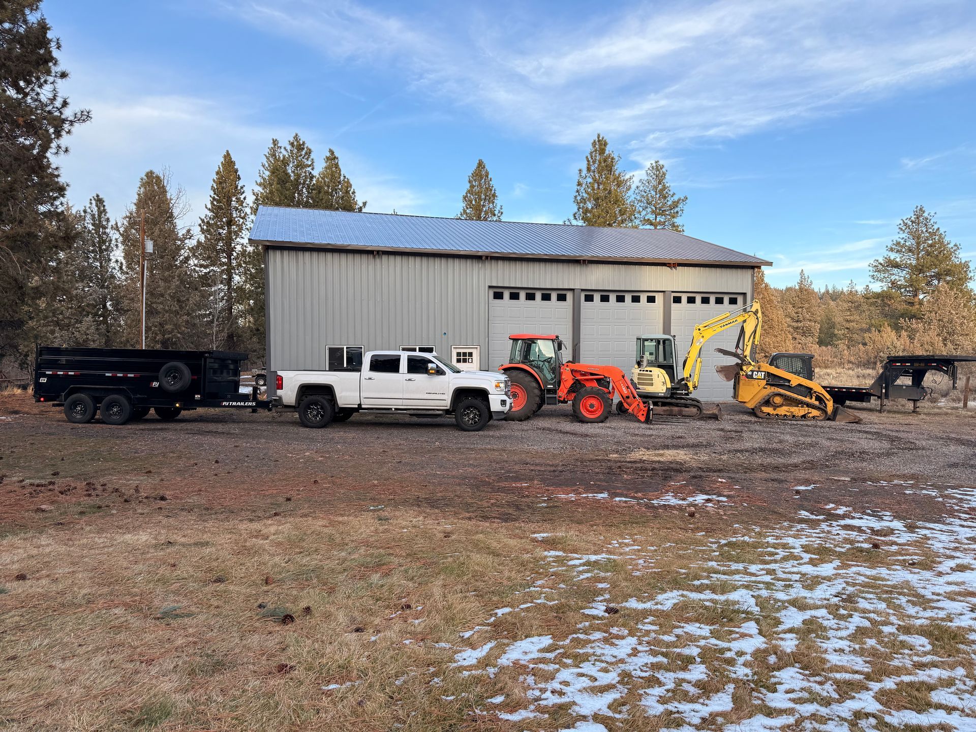 White trucks and construction equipment lined up outside a metal workshop on a snowy rural lot.