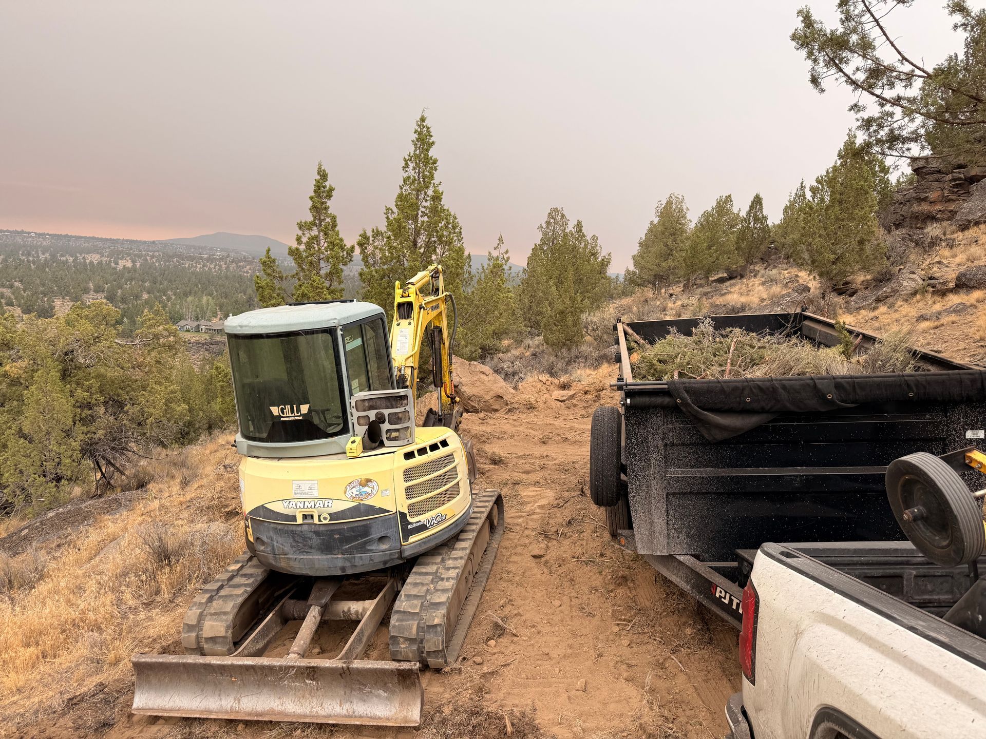 Yellow excavator on a dirt road beside a black dump trailer in a wooded hillside area
