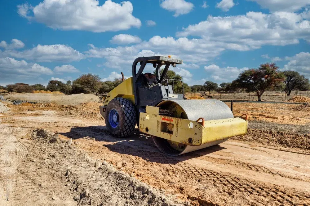 Yellow road roller on a dusty dirt road under a blue sky with clouds