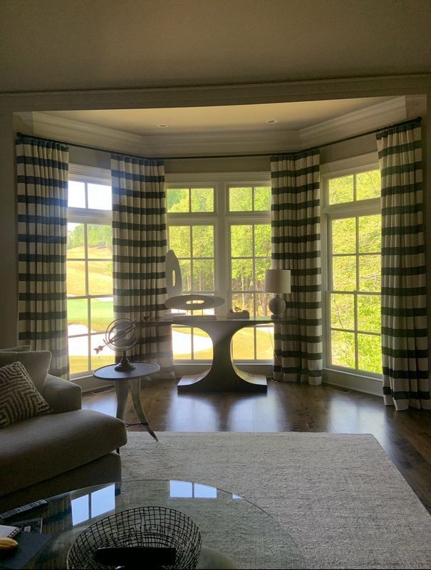 Sunlit living room with large bay windows, striped curtains, and a glass table by a sofa.