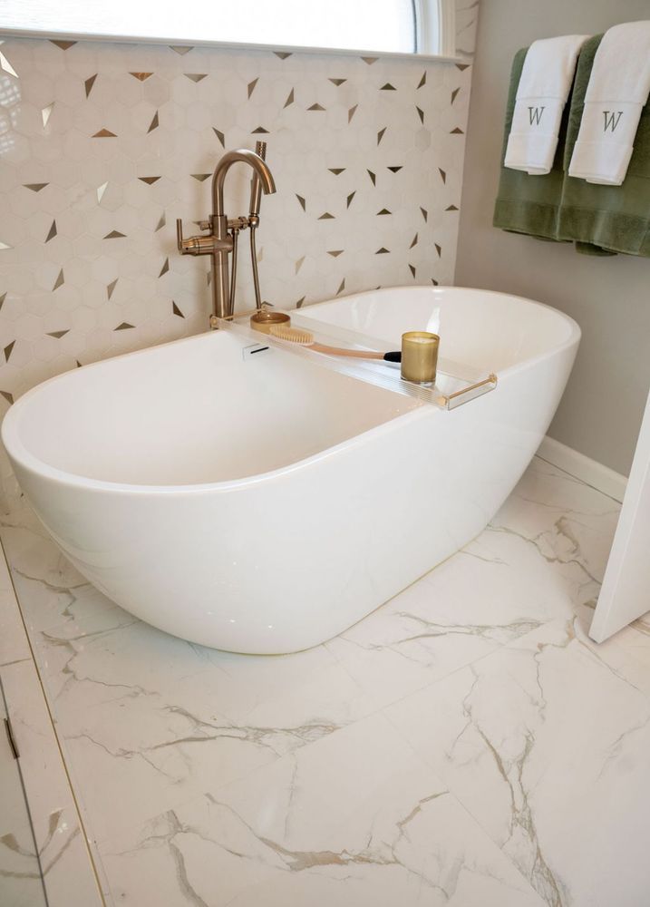 Modern freestanding white bathtub in a marble-tiled bathroom with brass faucet and tray with candle.