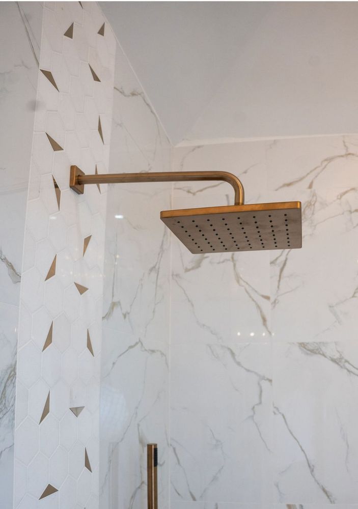 Brass rainfall showerhead on a white marble-tile shower wall with gold triangle accents