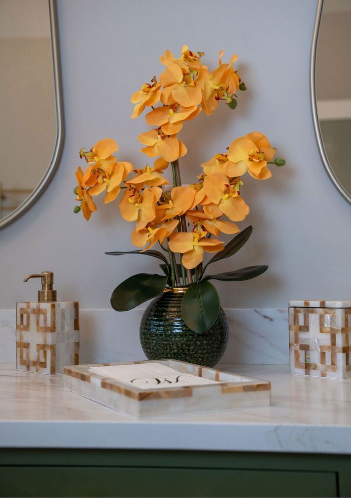 Yellow orchid arrangement in a green vase on a marble vanity with decorative soap dispensers and books