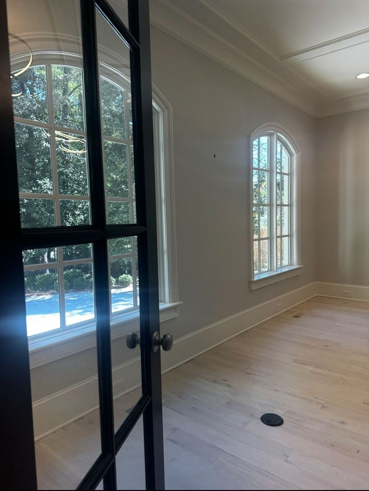 Bright empty room with white walls, light wood floors, arched window, and black-framed glass doors.