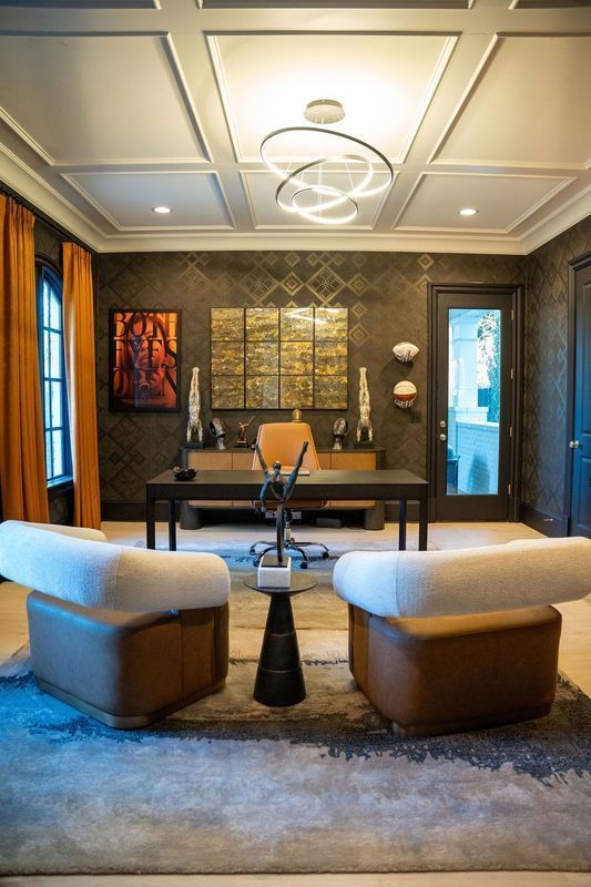 Modern office lounge with two cream chairs, dark wood walls, desk, and abstract art under a coffered ceiling