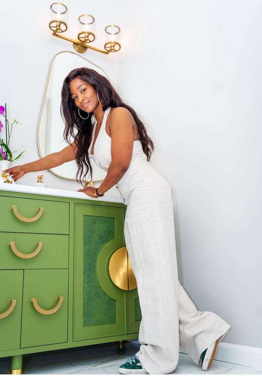 Woman in white leaning on a green dresser with gold handles and mirrors, in a bright room.