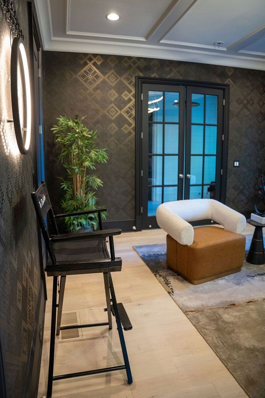 Modern living room with dark patterned walls, glass doors, black chairs, and a tan ottoman on a rug