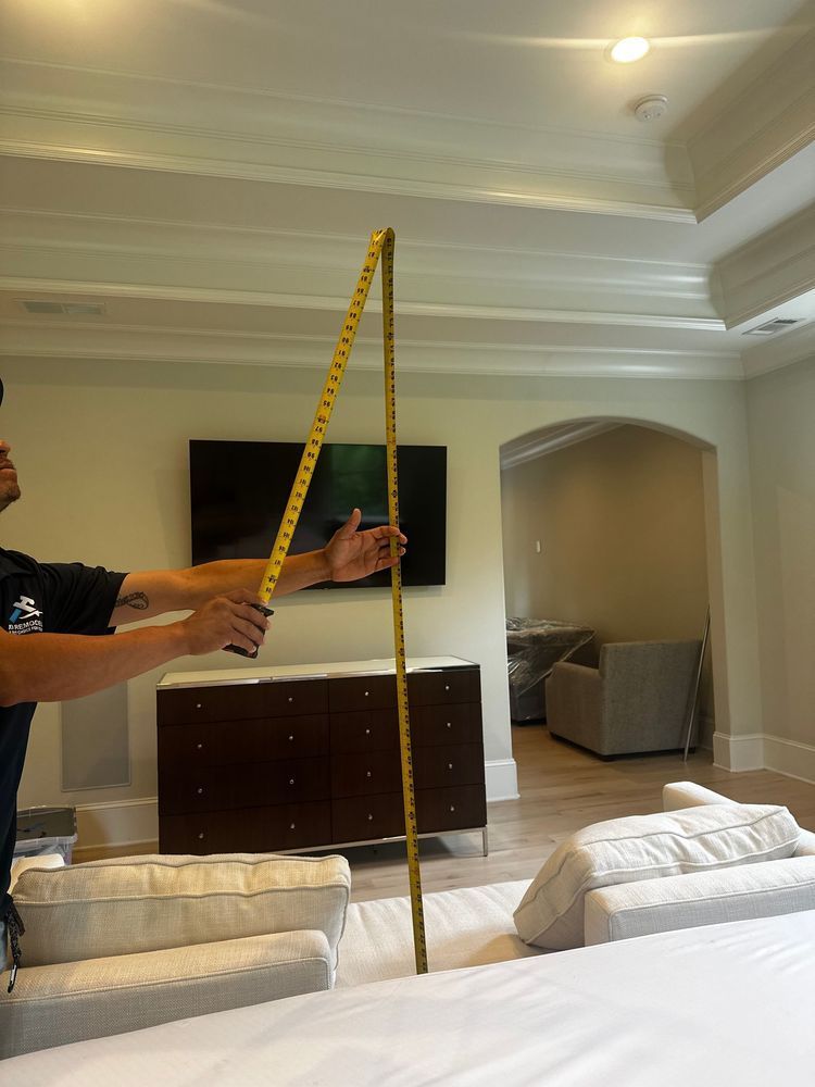 Hand holding a long yellow measuring tape in a bright living room with a sofa and dresser.