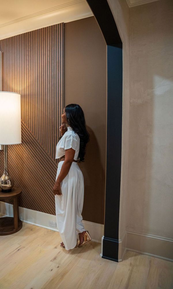 Woman in a white dress standing by a wall in a warmly lit room with striped curtains and a lamp.