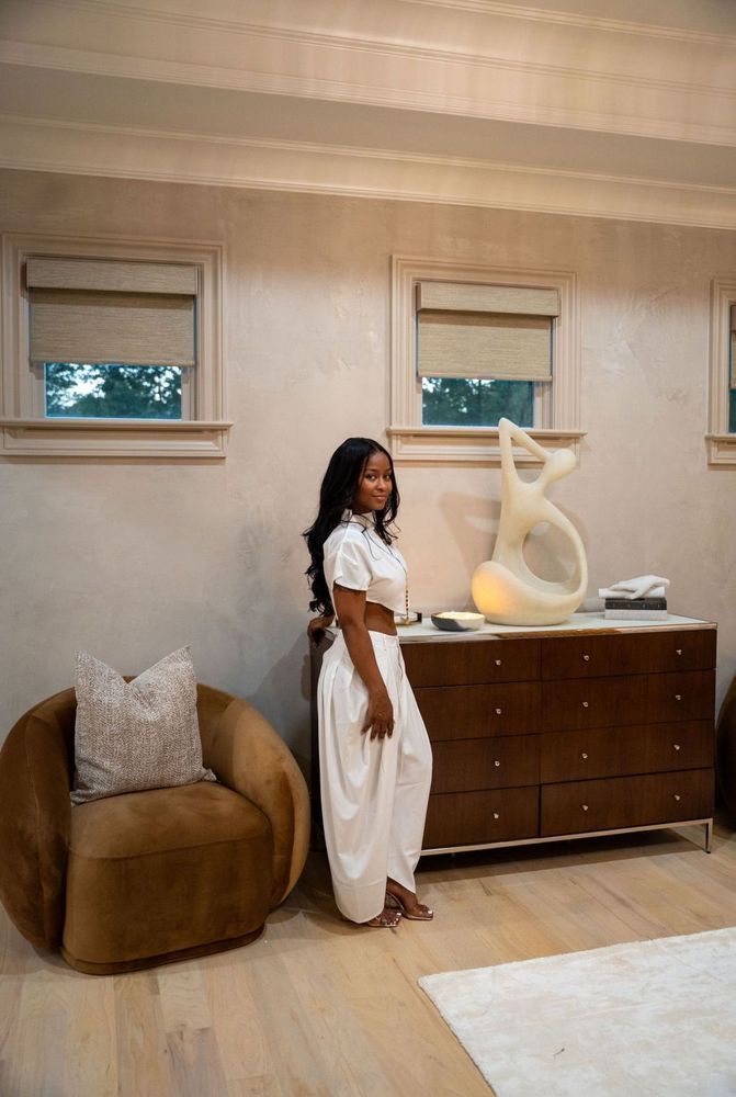 Woman standing in a modern bedroom beside a dresser, chair, and abstract sculpture