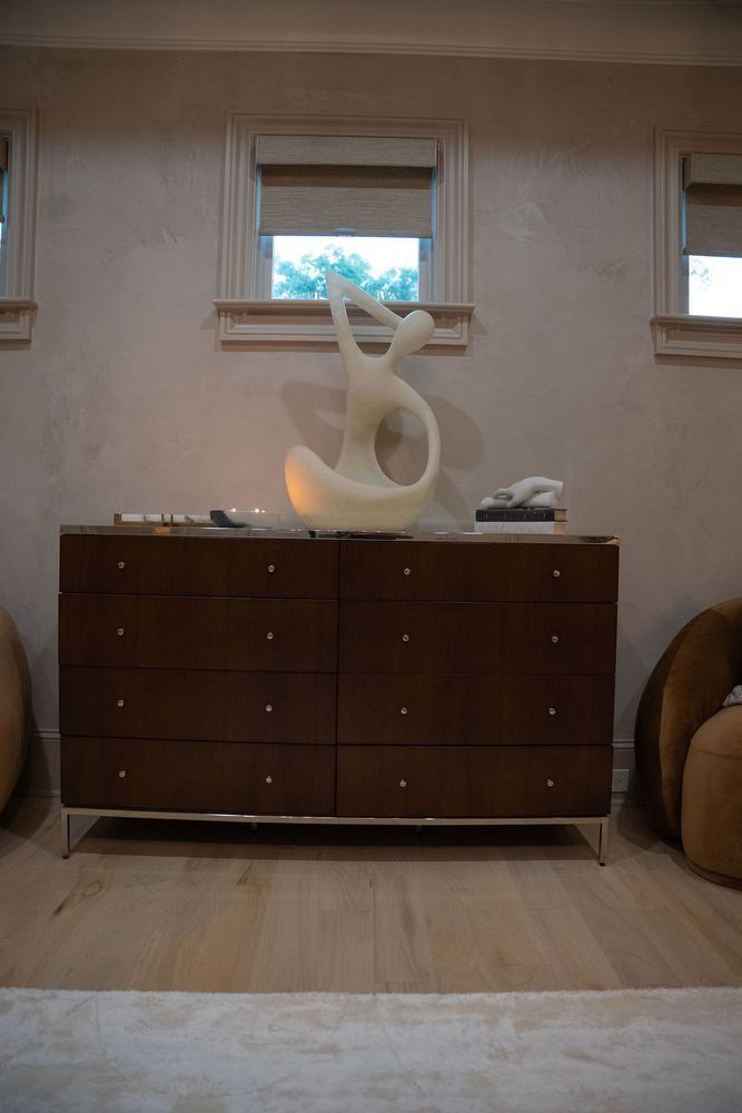 Abstract white sculpture on a dark wooden dresser in a softly lit room with windows.