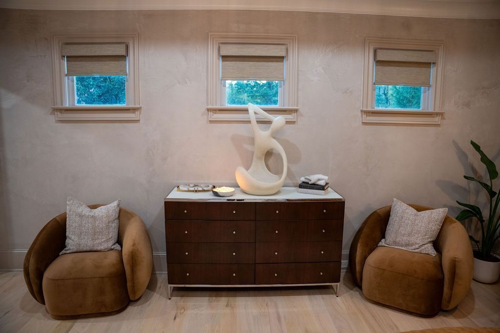 Modern living room with beige walls, three small windows, dark wood cabinet, and two brown chairs with pillows