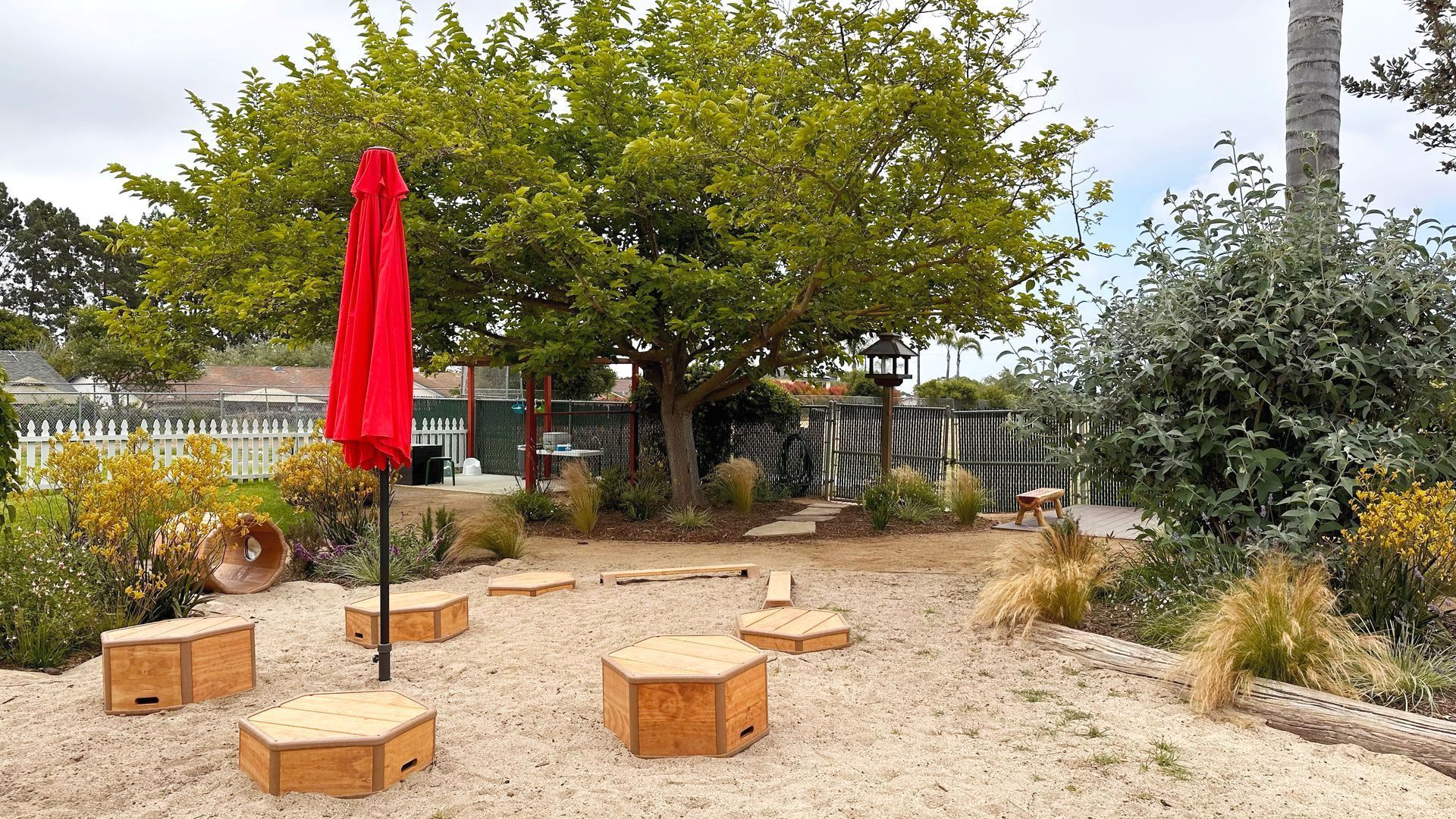 A child's play area with sand, wooden structures, and a red umbrella on a cloudy day.