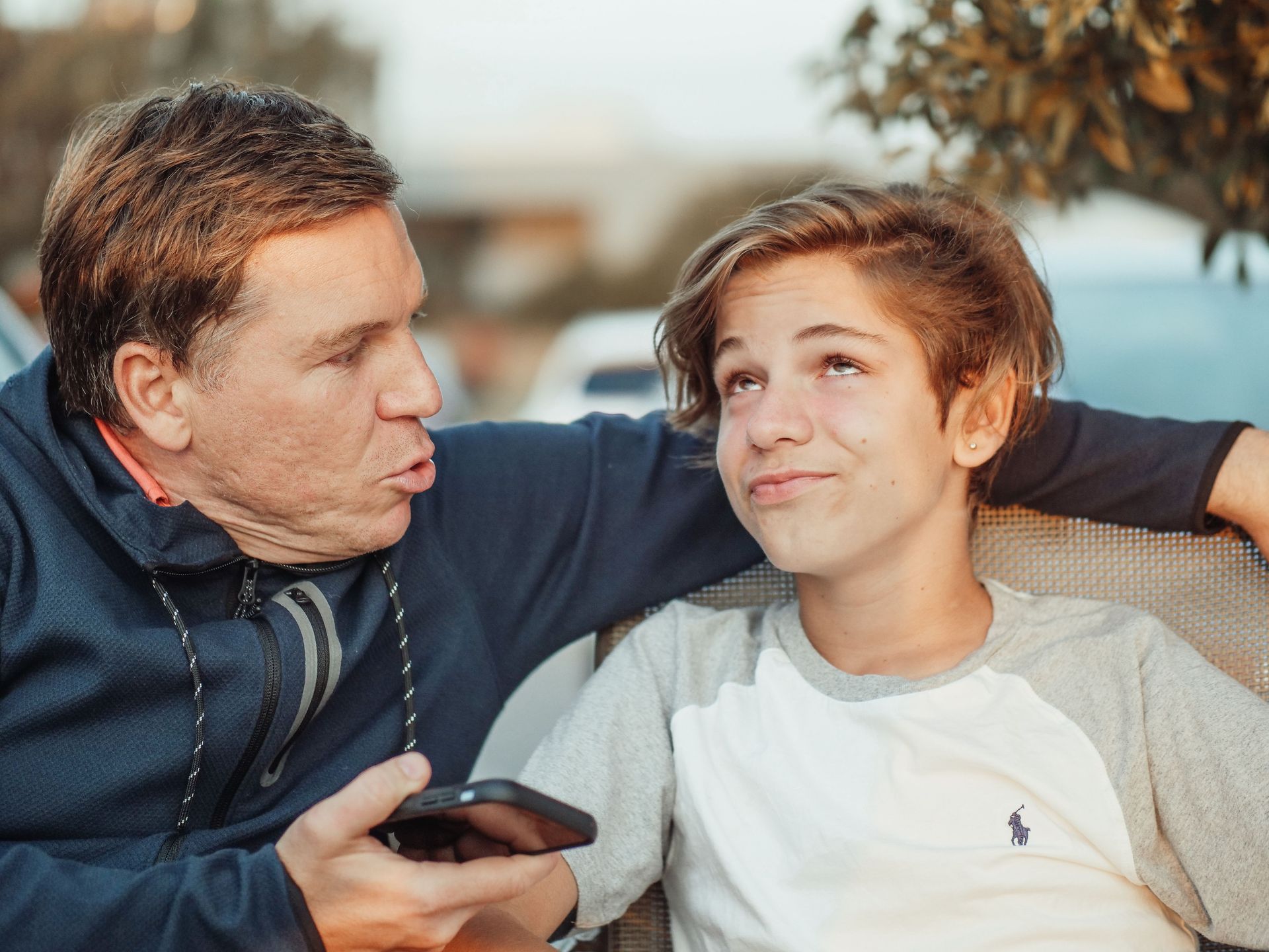Two people sitting on a couch, one speaking while holding a phone, the other smiling and looking up.