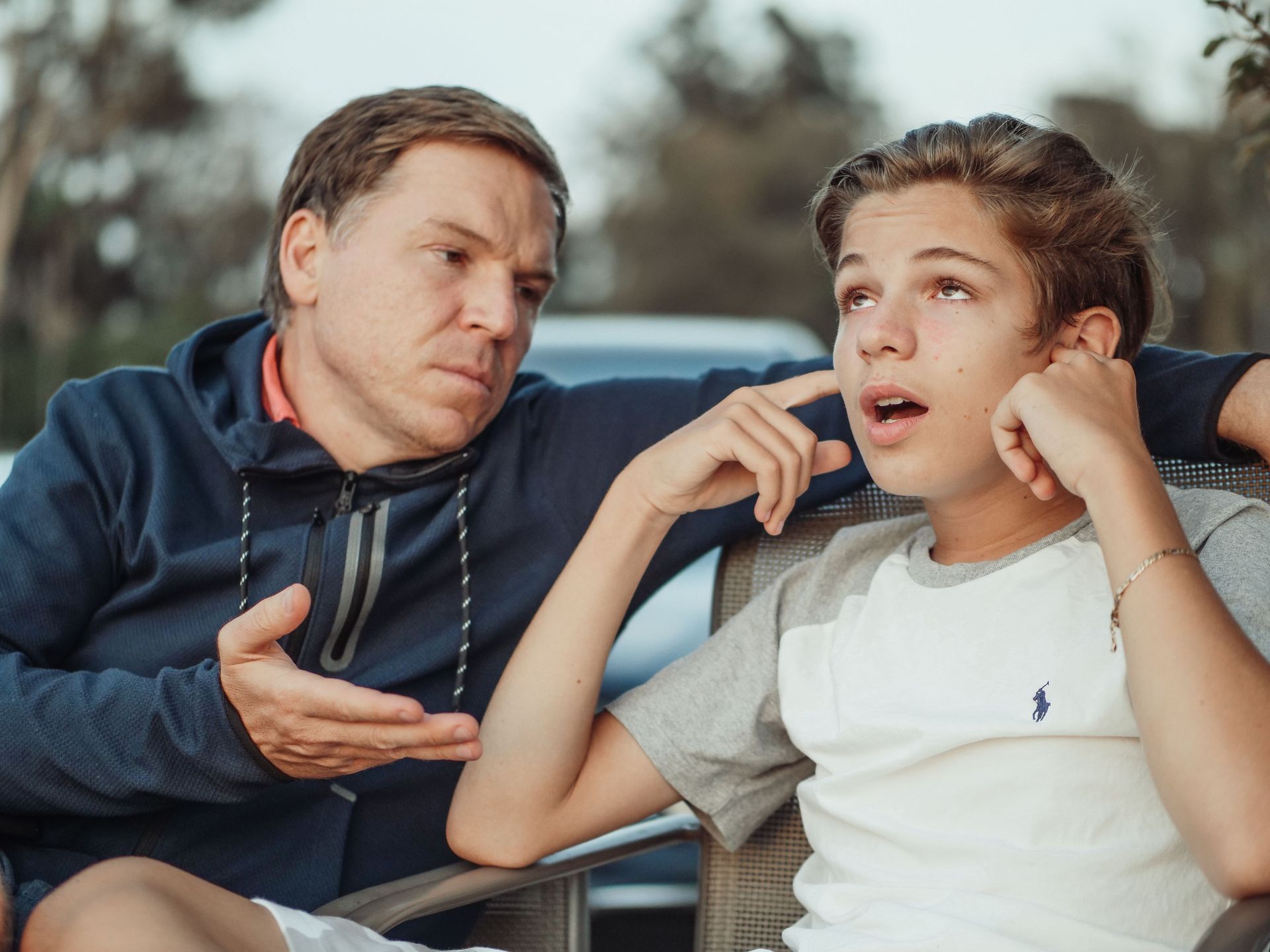 Two people on a couch talking, one in a navy hoodie and one in a white shirt, outdoors at dusk