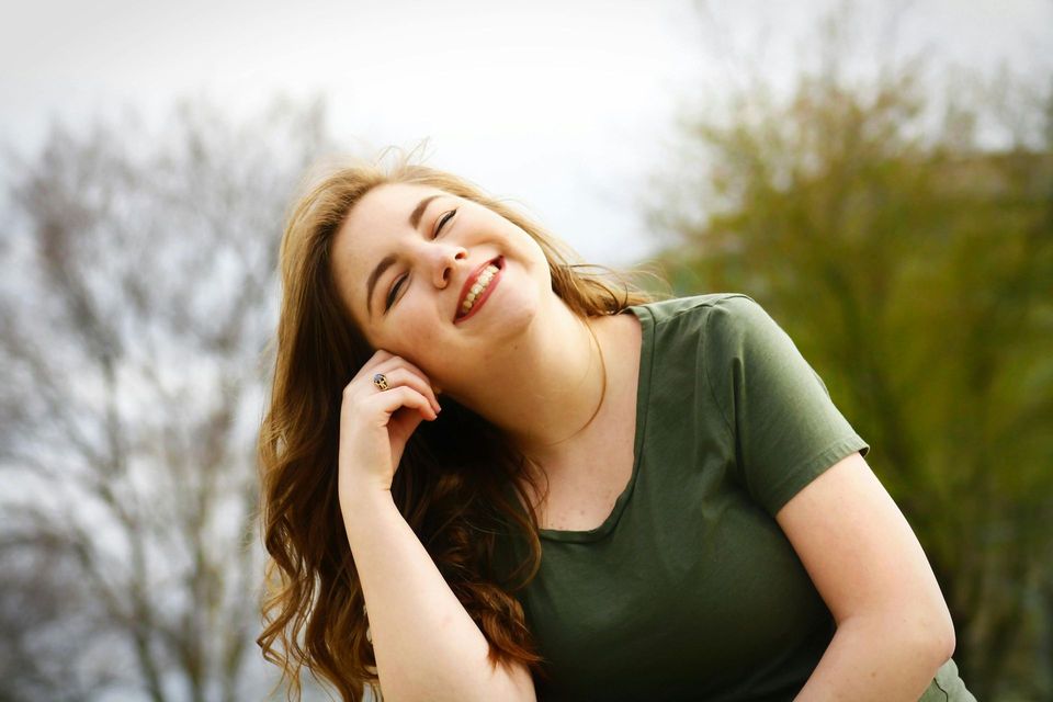 Una mujer con cabello largo y castaño sonríe, con la cabeza inclinada hacia atrás y vistiendo una camisa verde al aire libre.