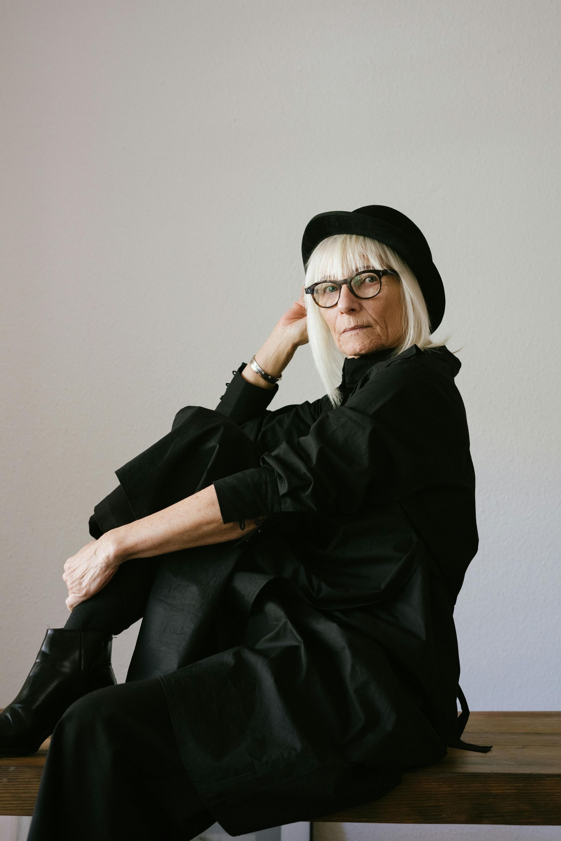 Person in black coat and hat sitting on a bench against a plain wall.