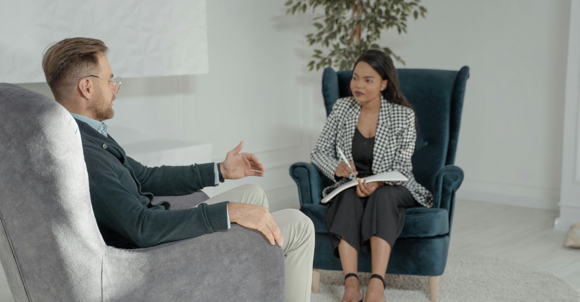 Two people in a counseling session seated in armchairs, one taking notes in a bright room.