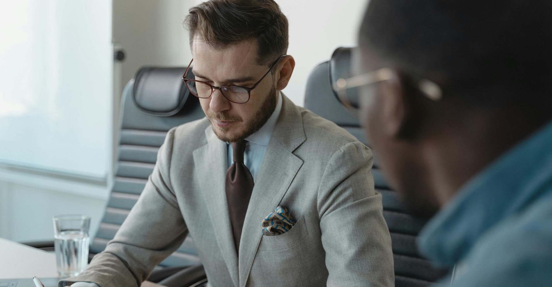 Two men in a conference room, one in a gray suit reviewing documents at a desk.