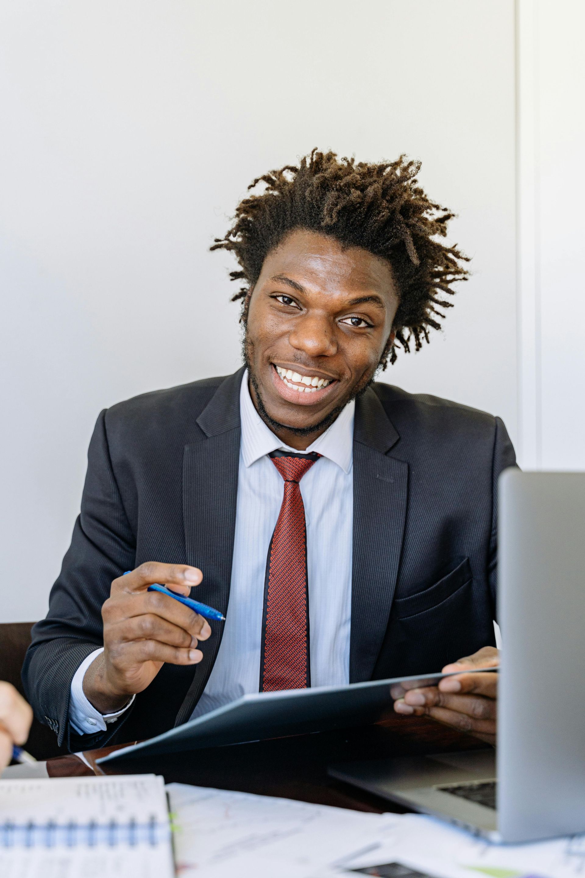 Businessperson in a suit smiling at a desk, holding a pen and reviewing notes beside a laptop.
