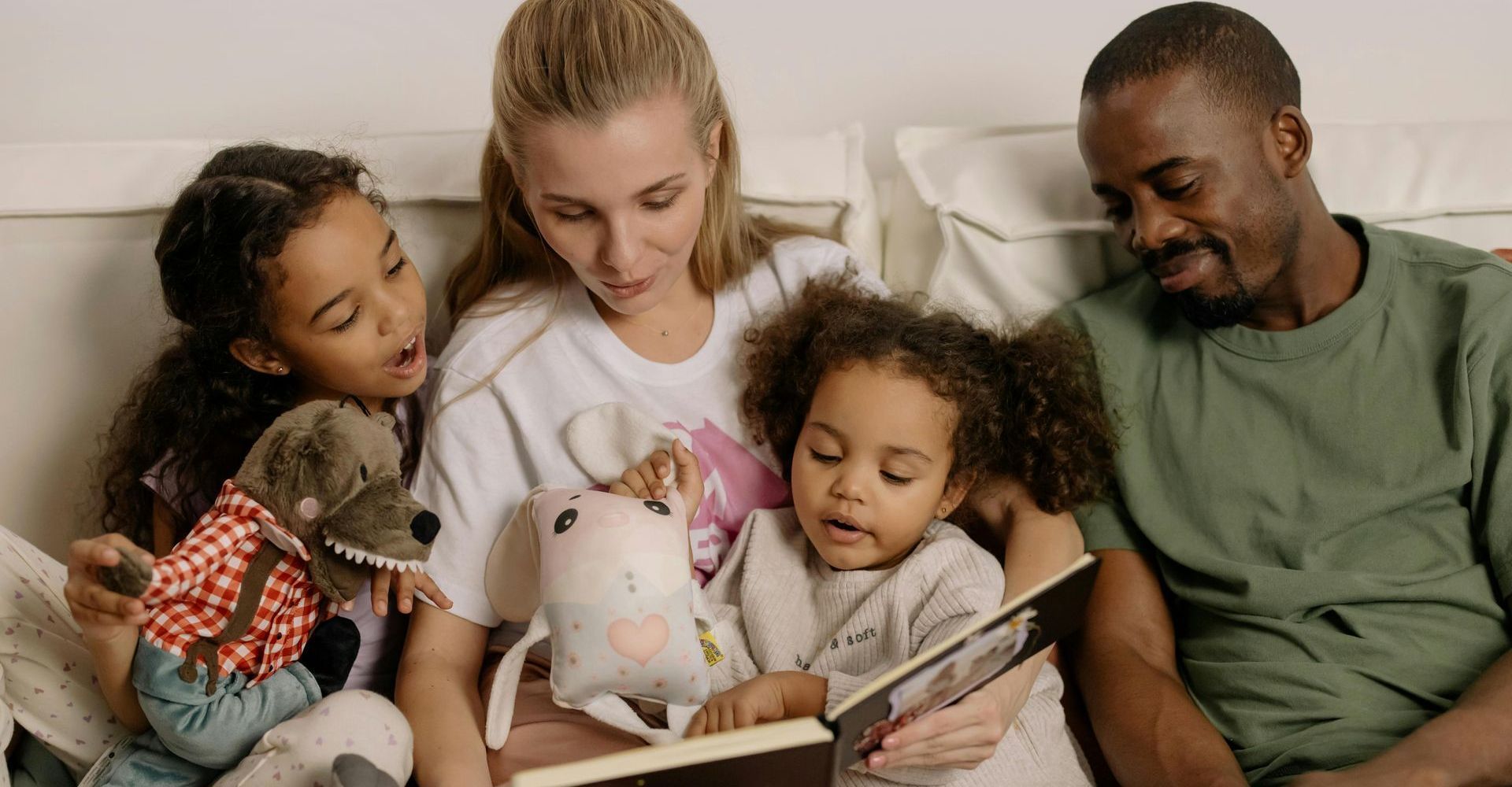 Family sitting on a couch reading a book together with a stuffed toy nearby