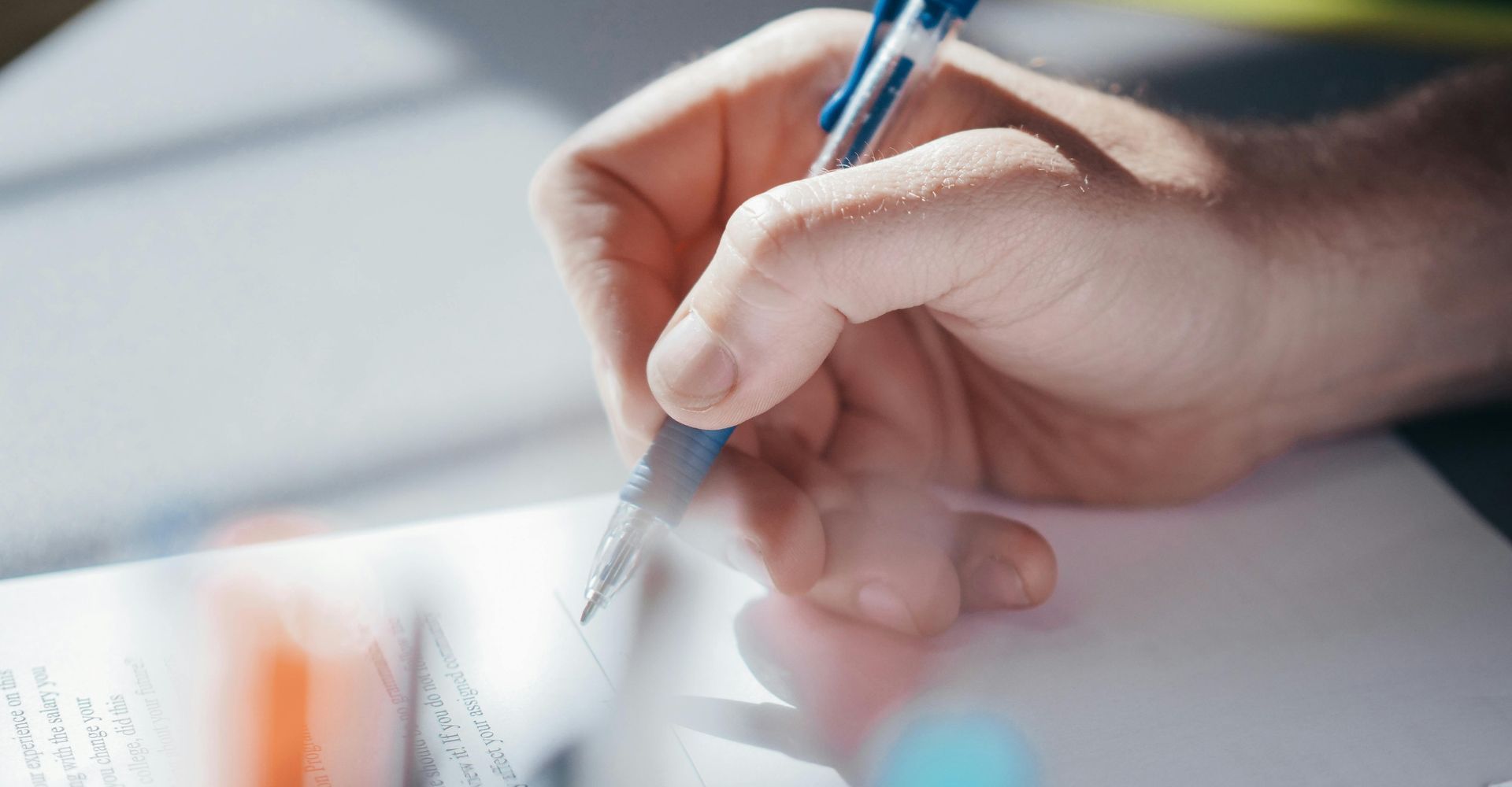 Hand holding a blue pen and writing on white paper near a bright window