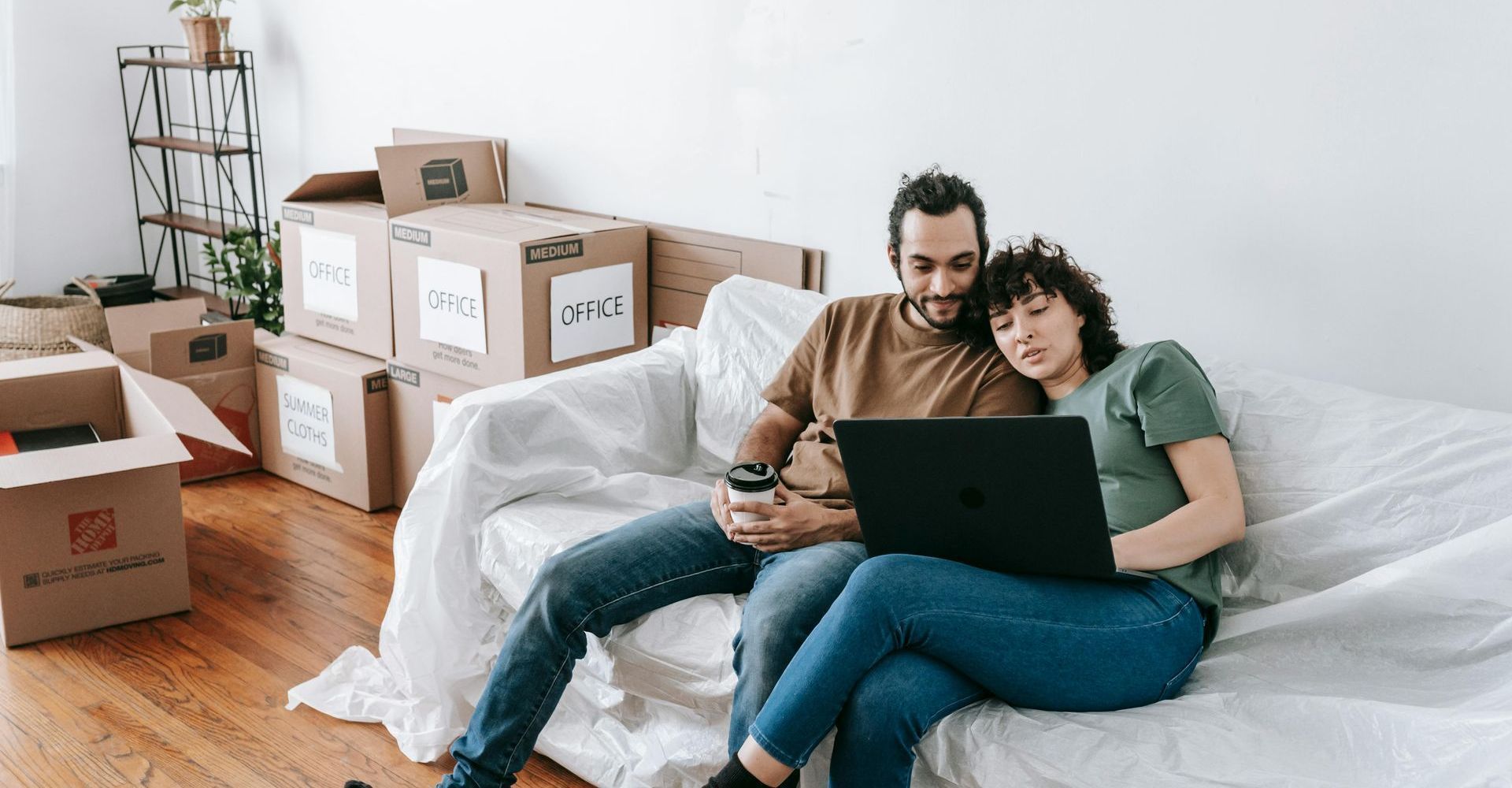 Couple sitting on a white couch using a laptop in a room with moving boxes
